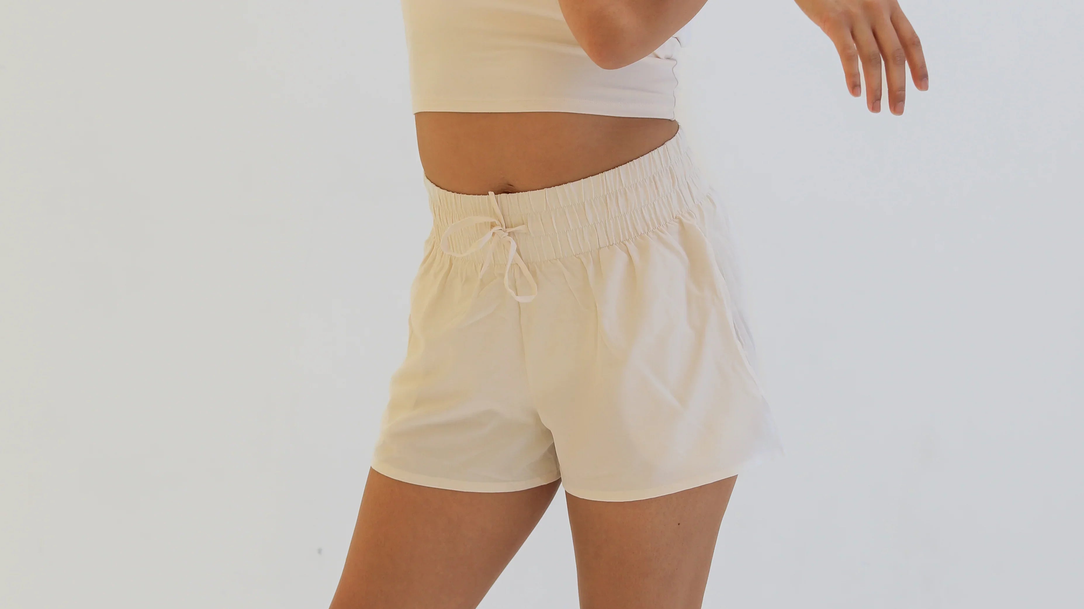 Full-body studio shot of a woman stretching her arms while wearing a cream-colored cropped tank top and matching loose-fitting, cream woven exercise shorts with a wide gathered elastic waistband.