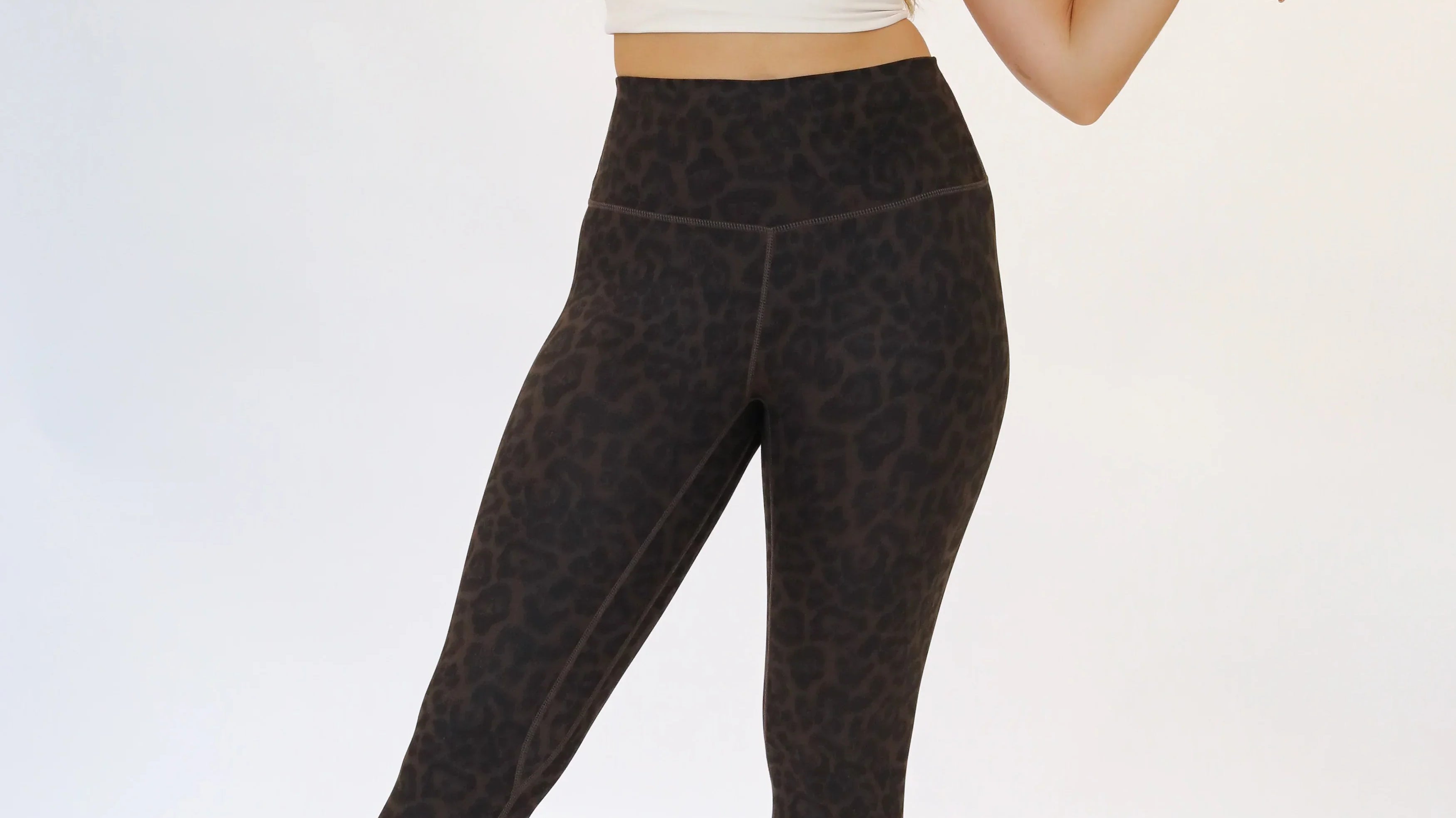 Full-body shot of a woman stretching her arms while wearing dark cocoa leopard print high-waisted leggings paired with a cream-colored cropped sports bra top and white athletic sneakers.