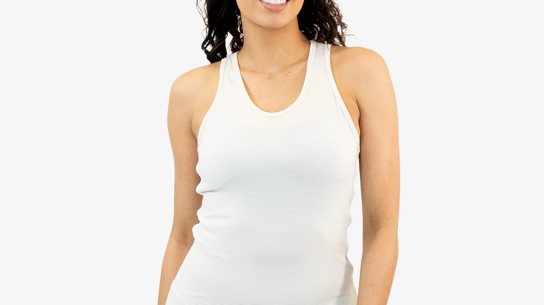 A smiling woman with curly hair wearing a white fitted racerback tank top paired with light sage green leggings against a plain light gray background.