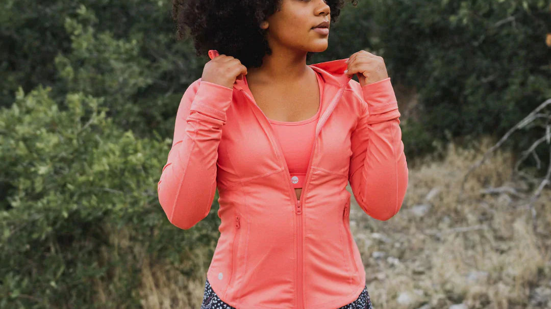 A woman with a dark curly afro looks off-camera while posing outdoors in a zippered coral pink performance jacket, a matching sports bra, and black patterned leggings.