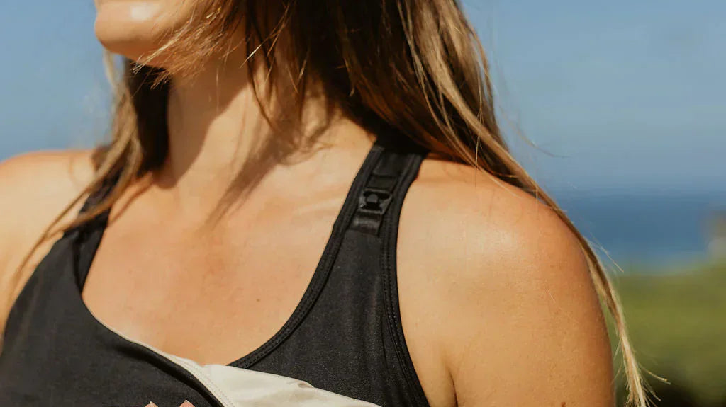 Outdoor lifestyle photo of a smiling woman wearing a black Senita maternity tank top and maroon bottoms, showing the easy-access drop-down nursing cup feature with a tan inner lining against a soft-focus blue sky background.