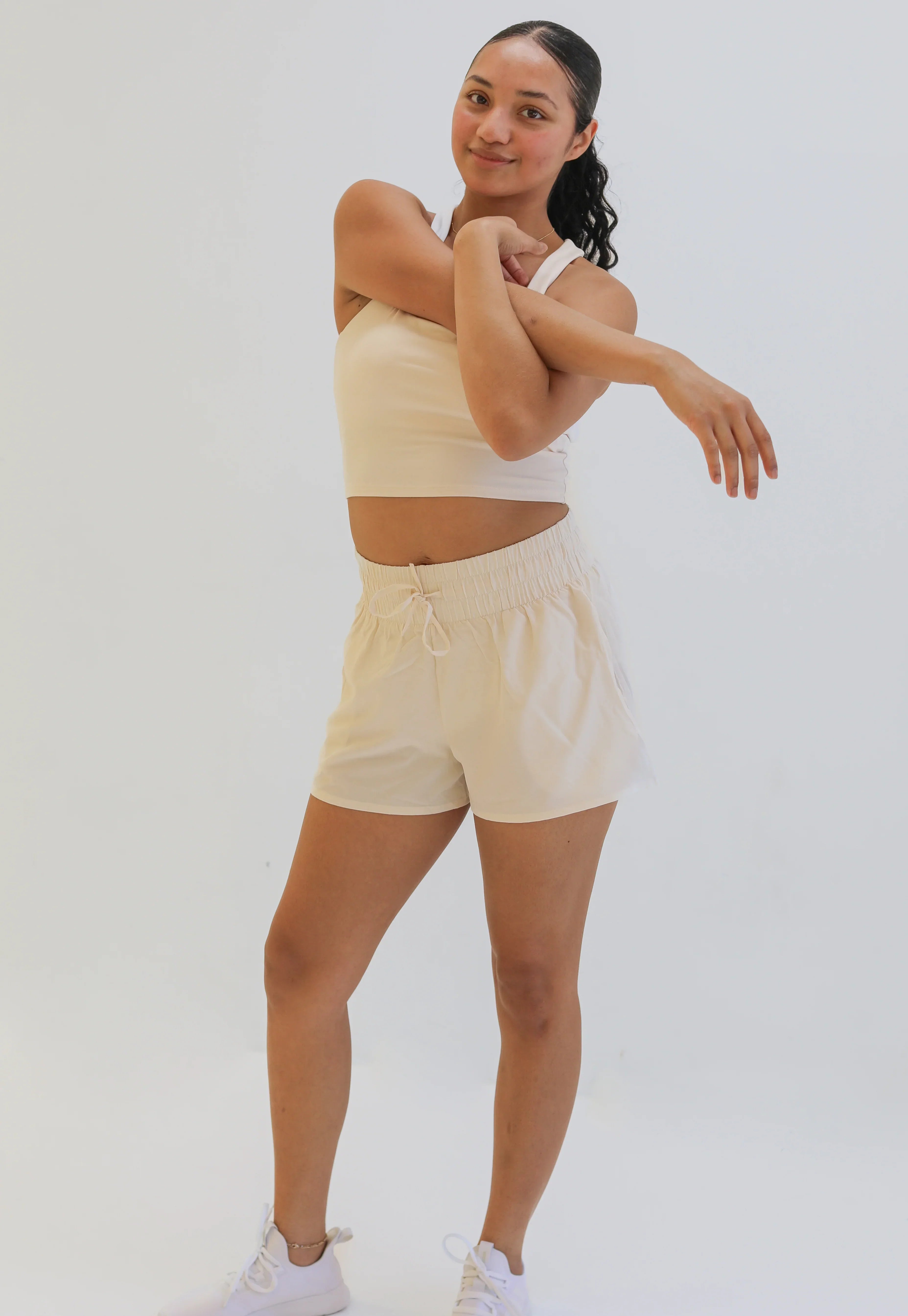 Full-body studio shot of a woman stretching her arms while wearing a cream-colored cropped tank top and matching loose-fitting, cream woven exercise shorts with a wide gathered elastic waistband.
