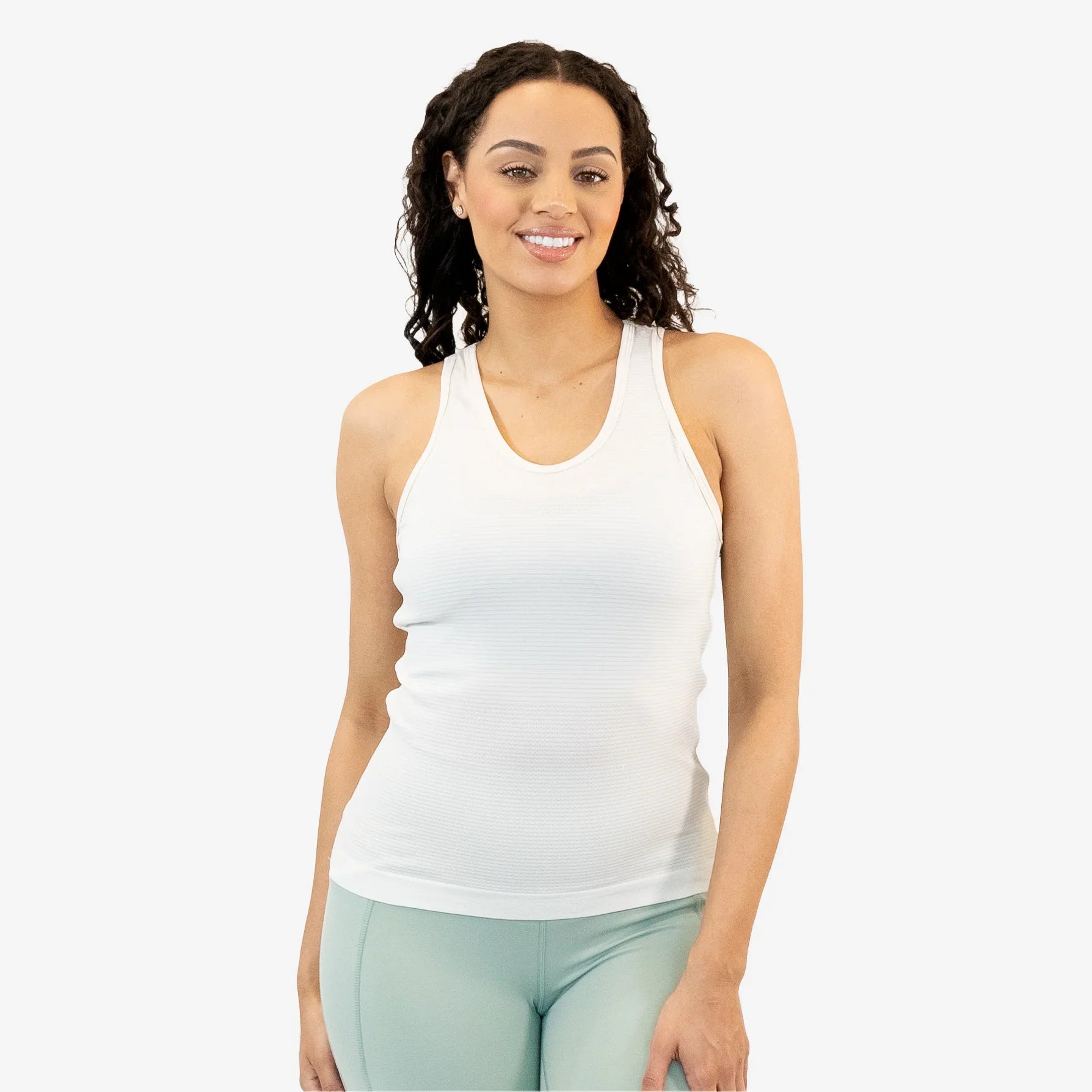 A smiling woman with curly hair wearing a white fitted racerback tank top paired with light sage green leggings against a plain light gray background.