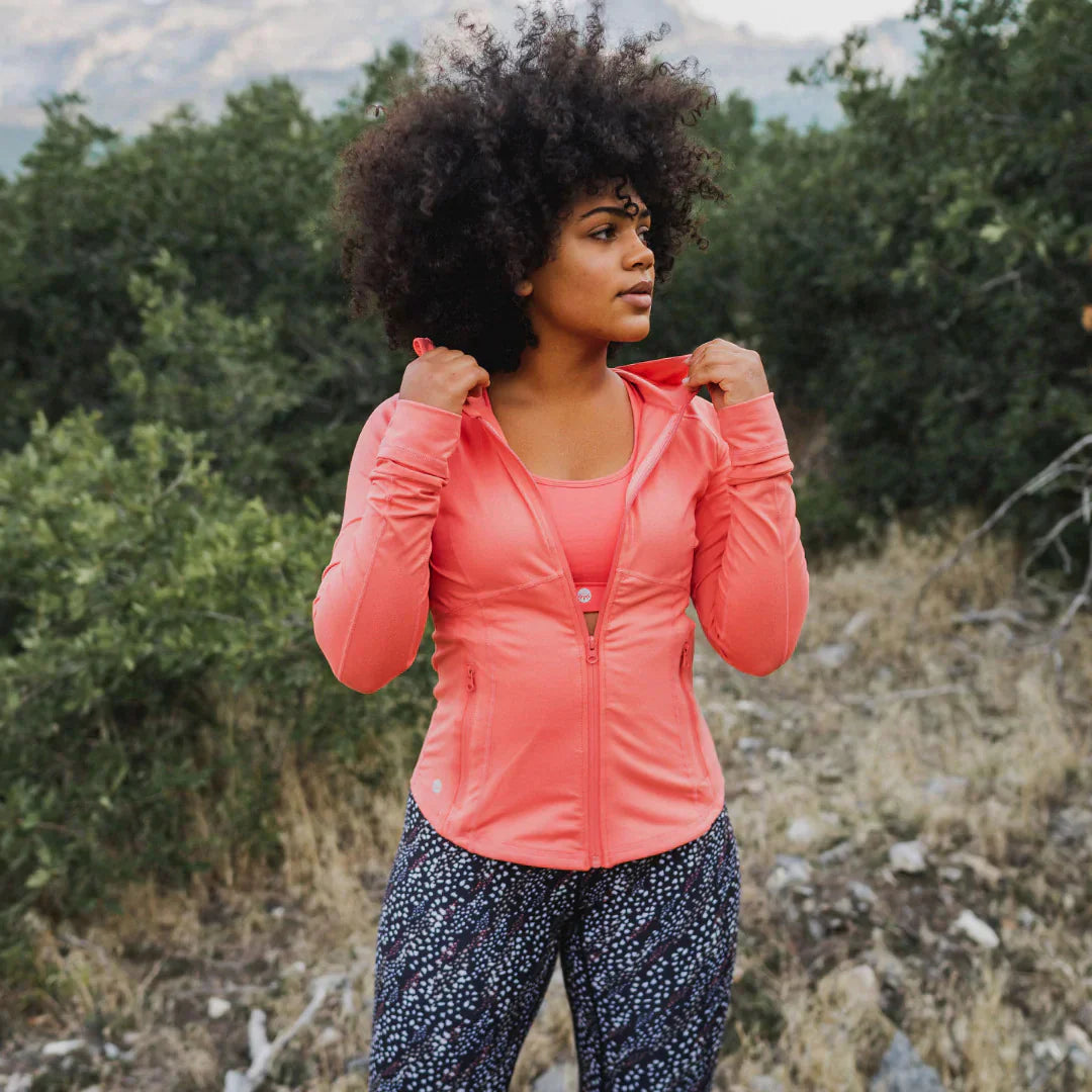 A woman with a dark curly afro looks off-camera while posing outdoors in a zippered coral pink performance jacket, a matching sports bra, and black patterned leggings.