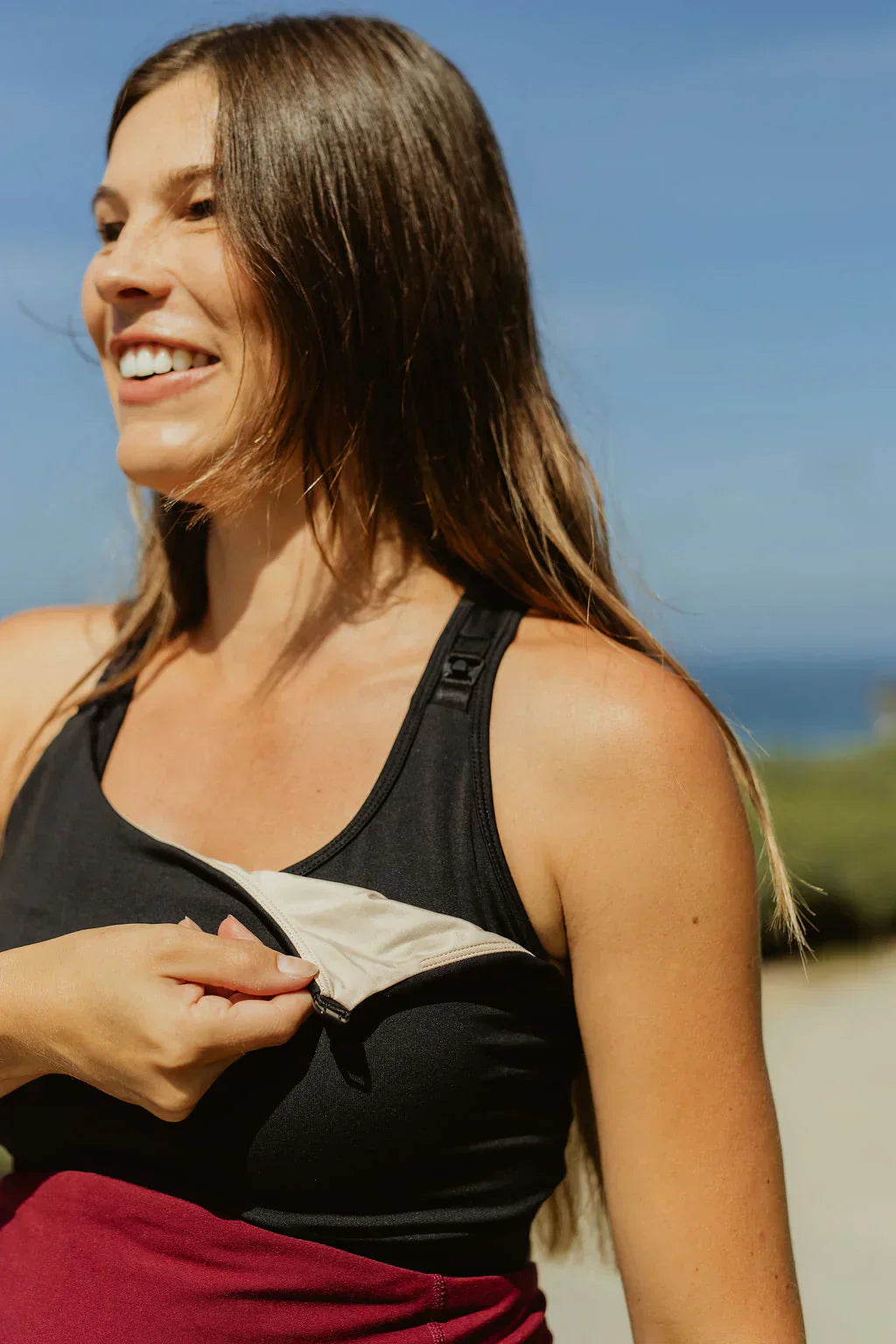 Outdoor lifestyle photo of a smiling woman wearing a black Senita maternity tank top and maroon bottoms, showing the easy-access drop-down nursing cup feature with a tan inner lining against a soft-focus blue sky background.
