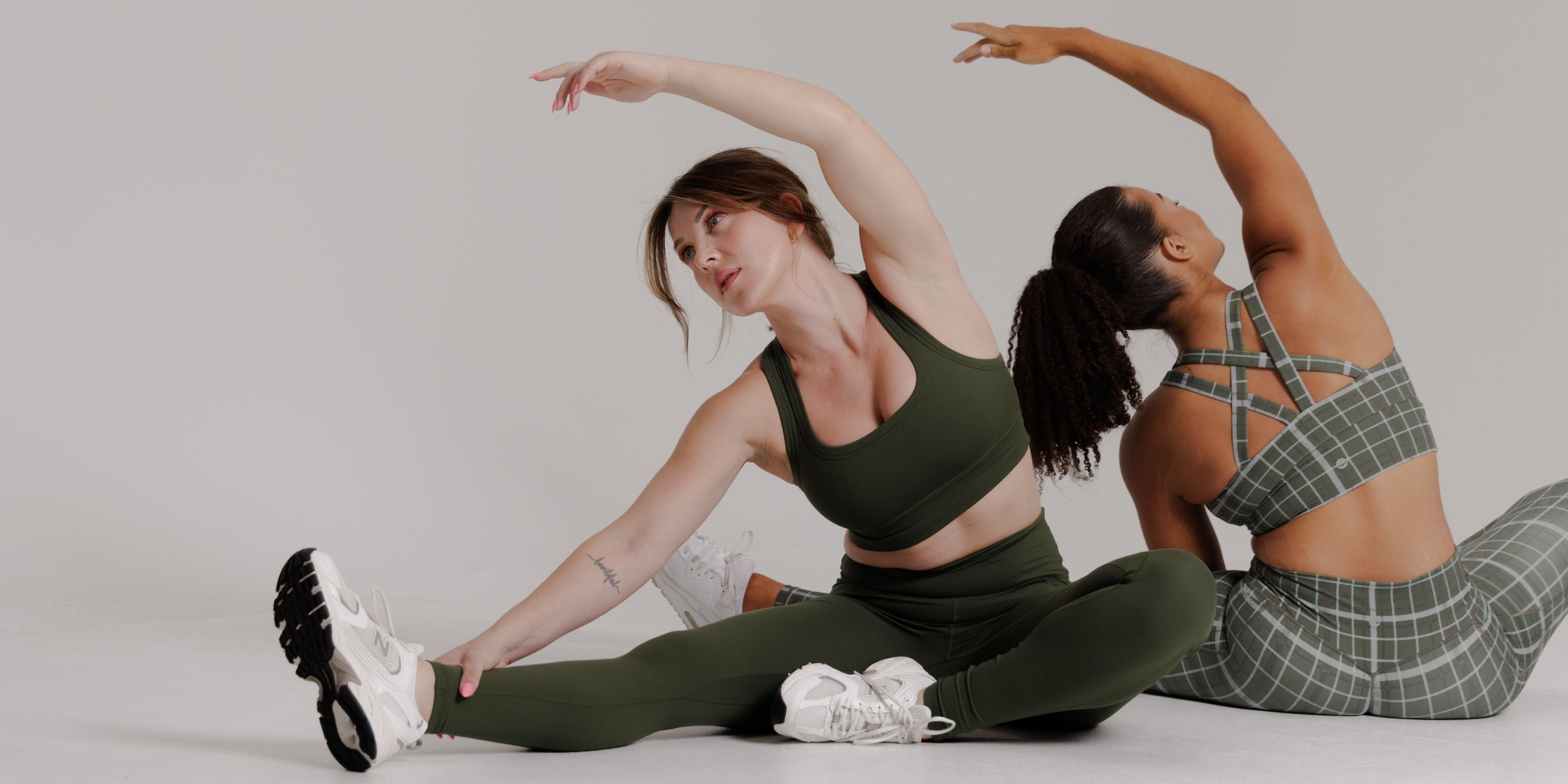Two women in matching athletic wear stretch side-by-side against a light gray studio background. The woman in front wears an olive green sports bra and leggings set with white sneakers, while the woman behind wears a green and gray plaid sports bra and matching plaid leggings.
