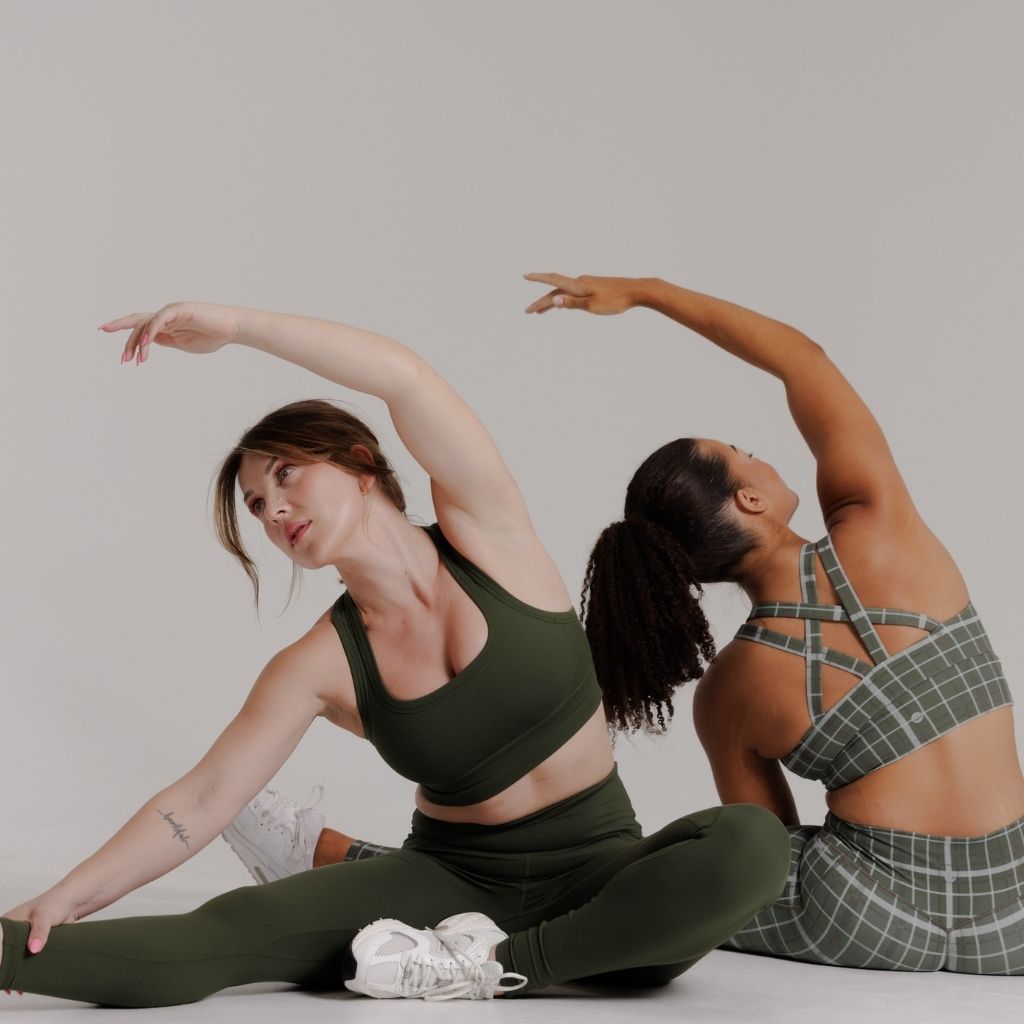 Two women in matching athletic wear pose together on a light gray studio floor. The woman in front wears an olive green sports bra and leggings set with white sneakers, while the woman behind wears a green plaid sports bra and matching plaid leggings.