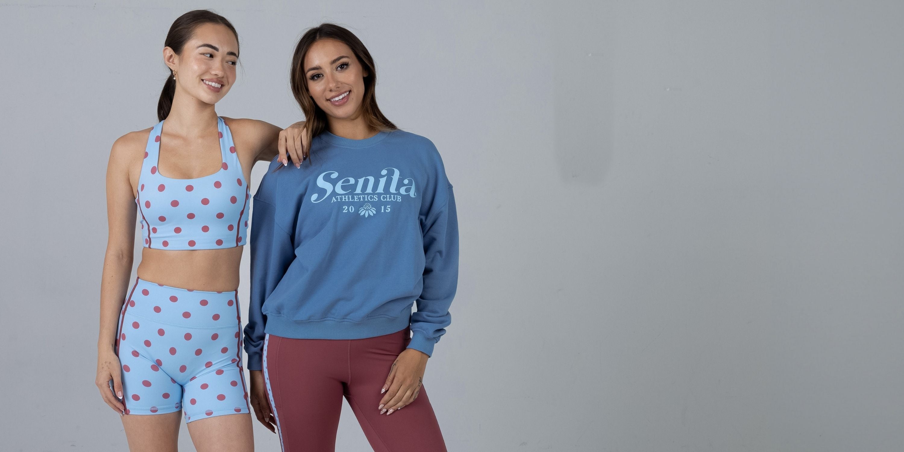 Two women modeling activewear from Senila Fitness. The woman on the left wears a light blue halter crop top and matching shorts with red polka dots.
