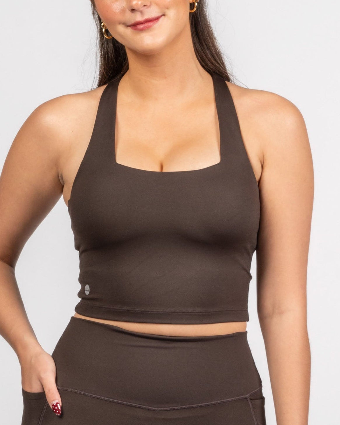 A woman modeling a matching brown activewear set consisting of a halter crop top and high-waisted leggings, photographed against a clean light gray studio background.